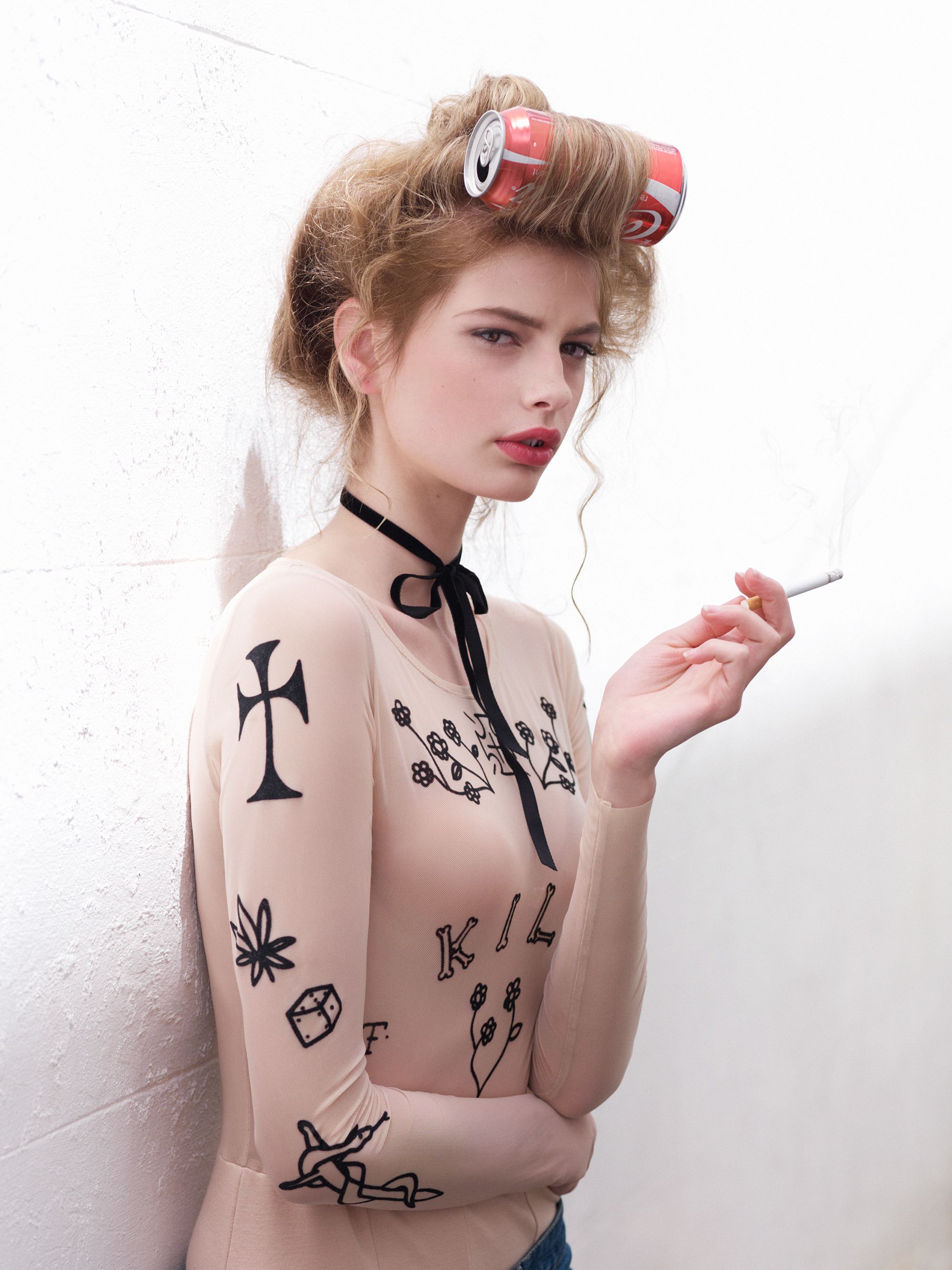 Woman in body suit with illustrated tattoo designs holding cigarette. She has soda can hair rollers and leans against a white wall.
