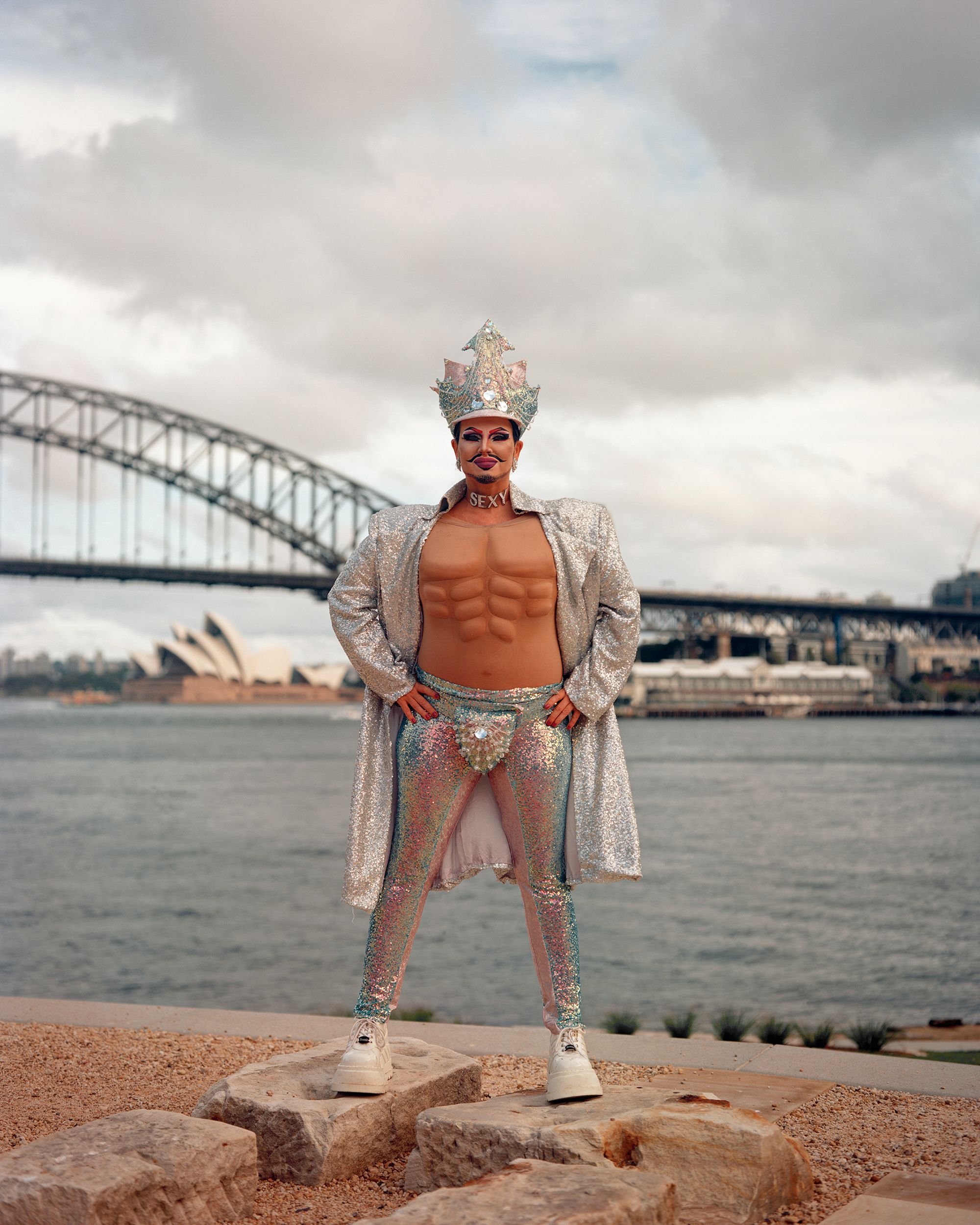 Drag queen poses in elaborate costume with Sydney Opera House and Harbour Bridge in background. Outdoor performance portrait.