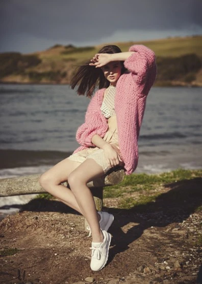 A young woman sits on a wooden rail near the water, wearing a pink cardigan, white top, and shorts, shielding her eyes from the sun.