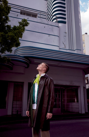 A man in a brown overcoat looks up at the striking Art Deco architecture of a multi-story building with white and blue stripes.