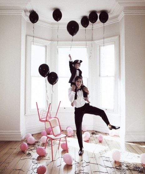 Man carries child on shoulders amidst balloons in a bright room, creating a festive and playful scene with party decorations and casual wear.