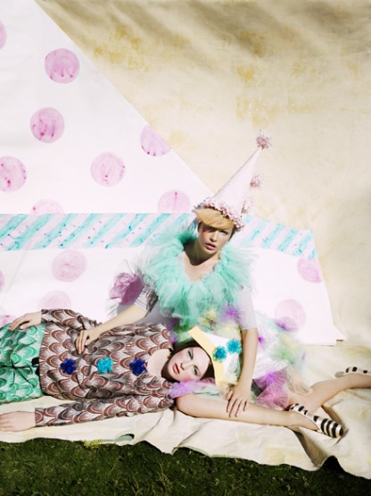 Two women in elaborate vintage-style costumes with pastel colors, posed on a blanket in an outdoor setting with theatrical, whimsical vibe.