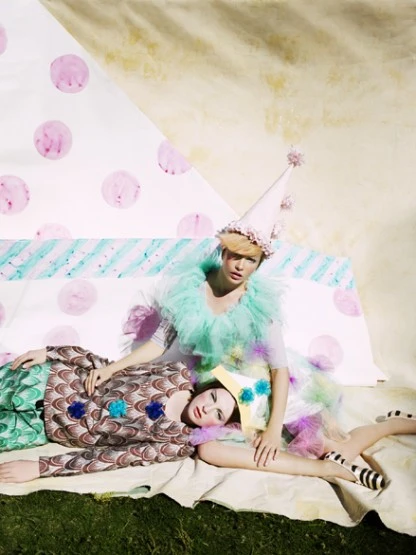 Two women in elaborate vintage-style costumes with pastel colors, posed on a blanket in an outdoor setting with theatrical, whimsical vibe.