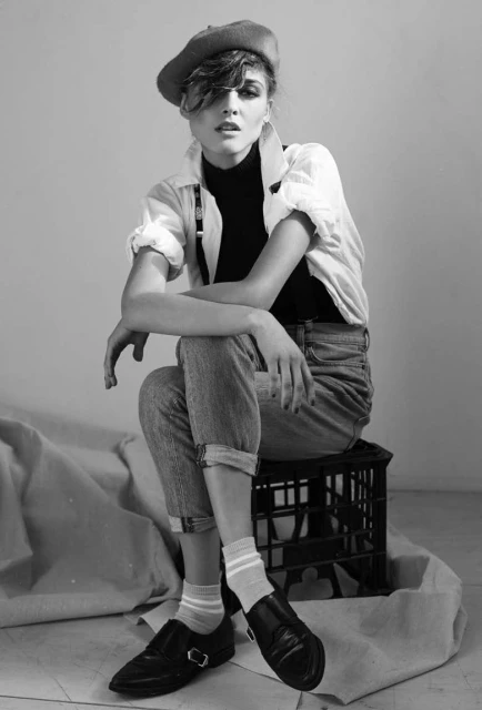 Monochrome studio portrait of a woman in a beret, jeans, and suspenders sitting on a crate, creating a vintage fashion vibe.