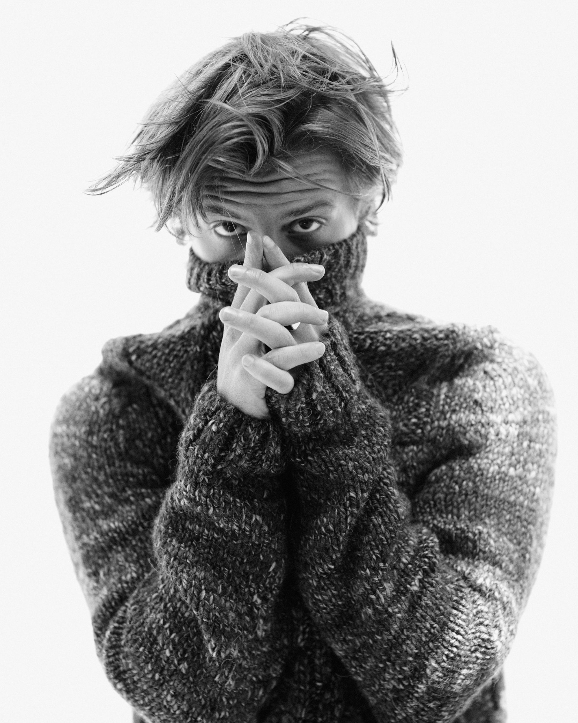 Black and white portrait of a young adult male wearing a knitted turtleneck sweater, hands clasped at his chin, with soft studio lighting.