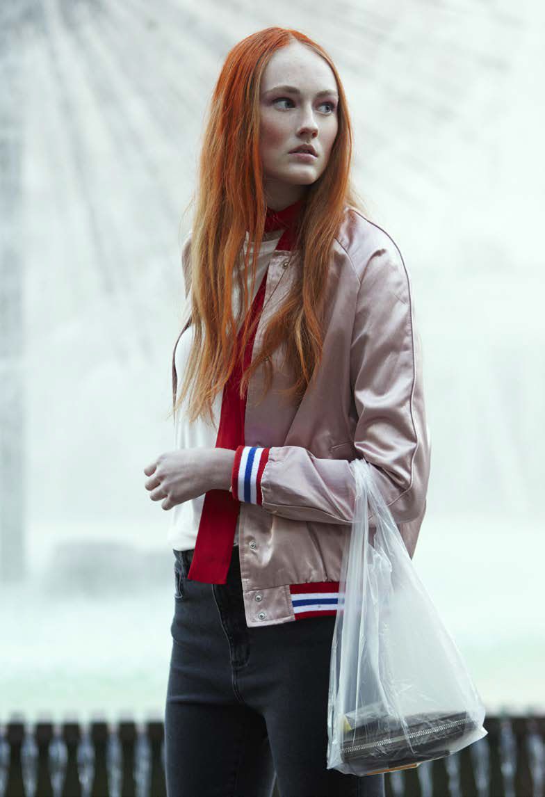 Woman with long red hair wearing a pink bomber jacket and holding a plastic shopping bag. She stands in front of a fountain.