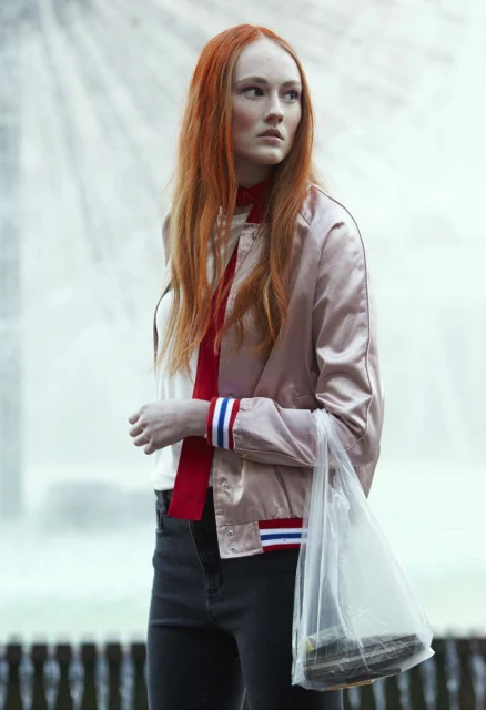Woman with long red hair wearing a pink bomber jacket and holding a plastic shopping bag. She stands in front of a fountain.