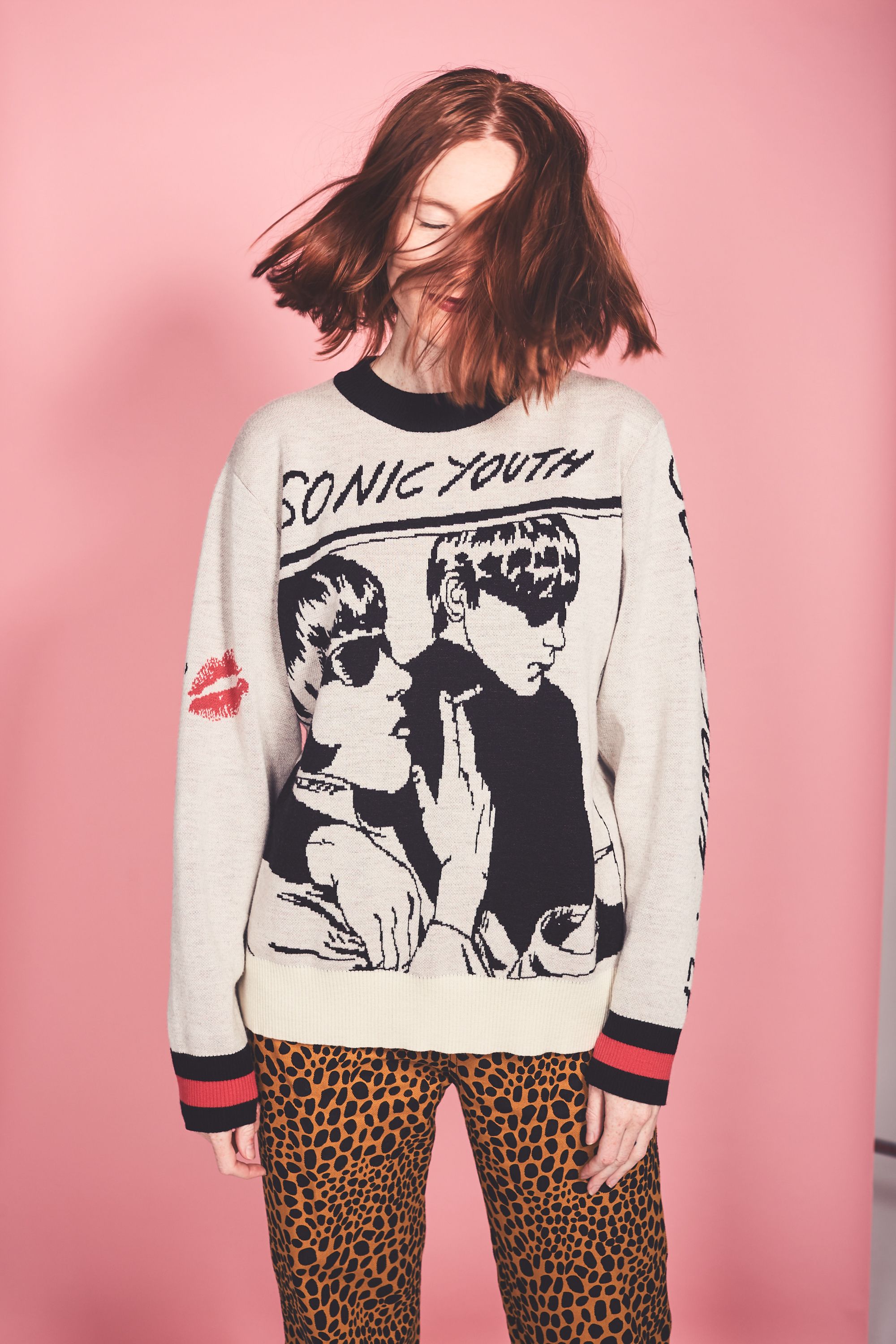 Young woman with red hair wearing a Sonic Youth graphic sweater and leopard-print pants against a pink background in a studio shot.