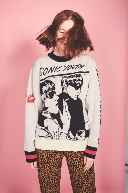 Young woman with red hair wearing a Sonic Youth graphic sweater and leopard-print pants against a pink background in a studio shot.