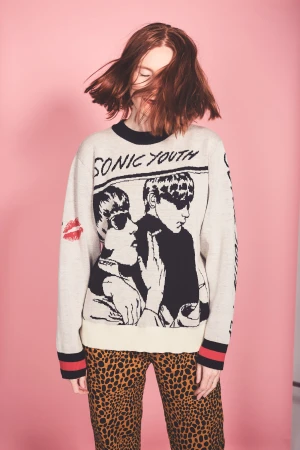 Young woman with red hair wearing a Sonic Youth graphic sweater and leopard-print pants against a pink background in a studio shot.