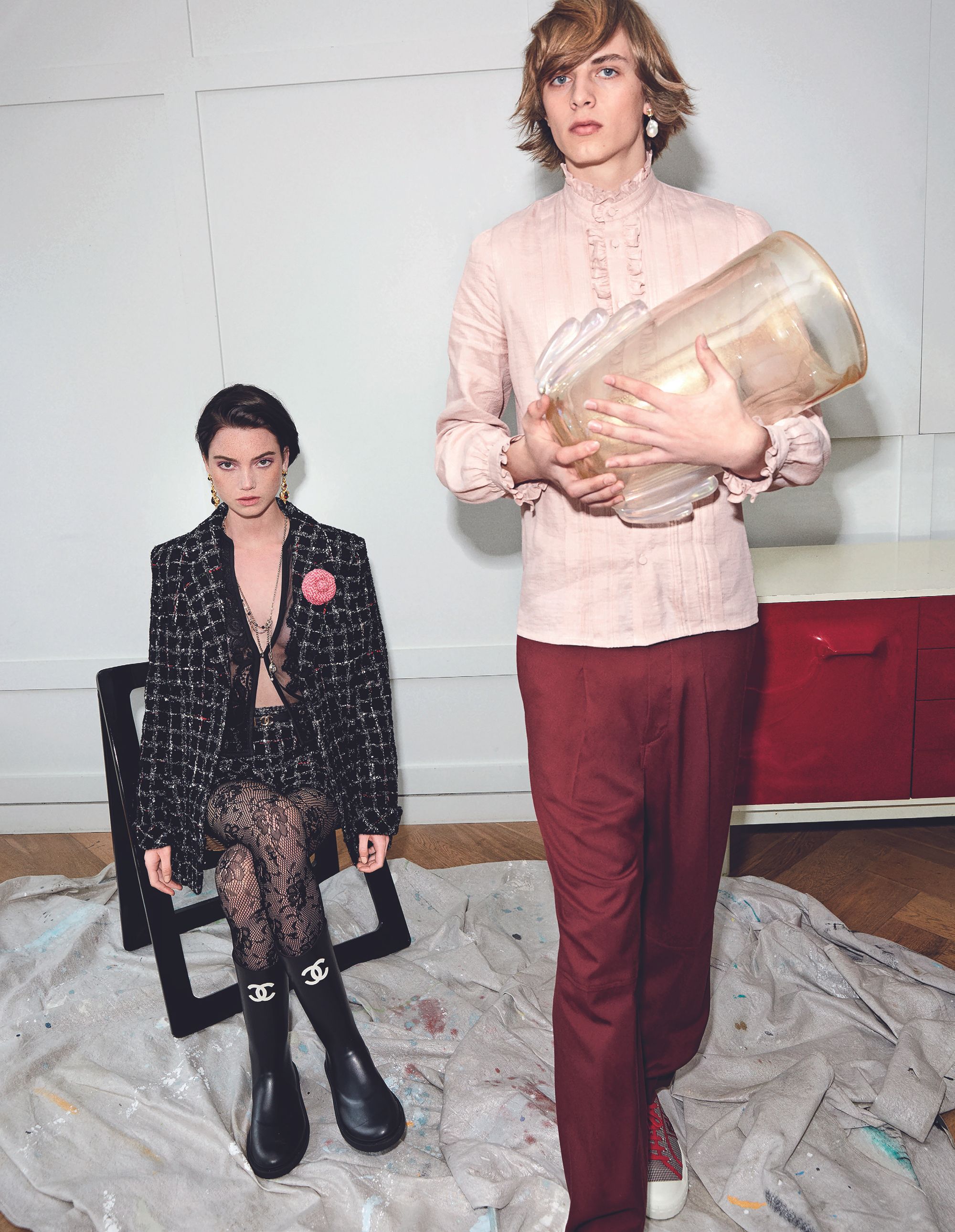 Two fashion models, one seated wearing tweed suit with Chanel boots, the other standing holding glass vase, in a modern interior.