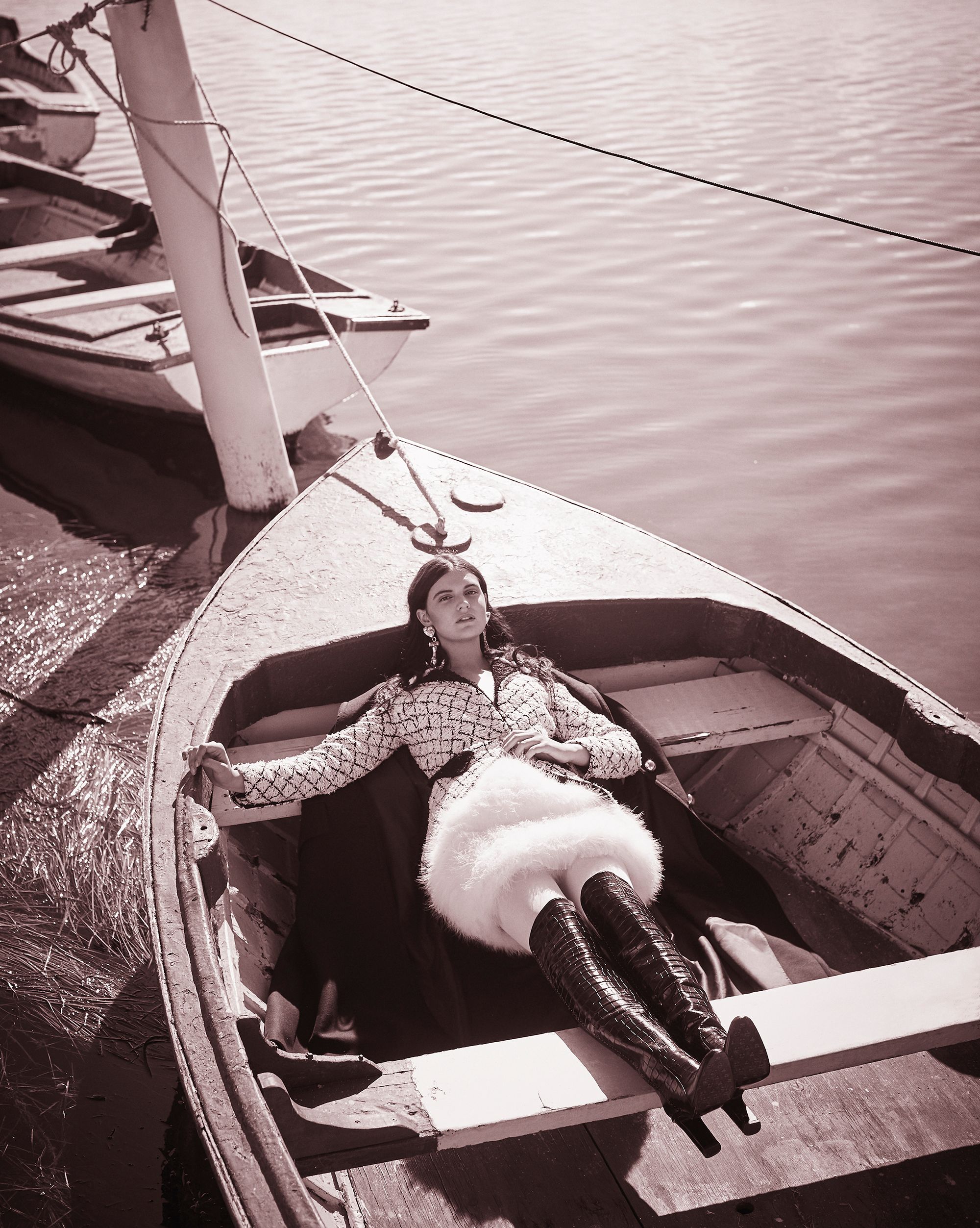 A fashionable woman relaxes in a small rowboat on the water, wearing a patterned coat, short furry skirt, and tall boots. Several other boats are nearby.