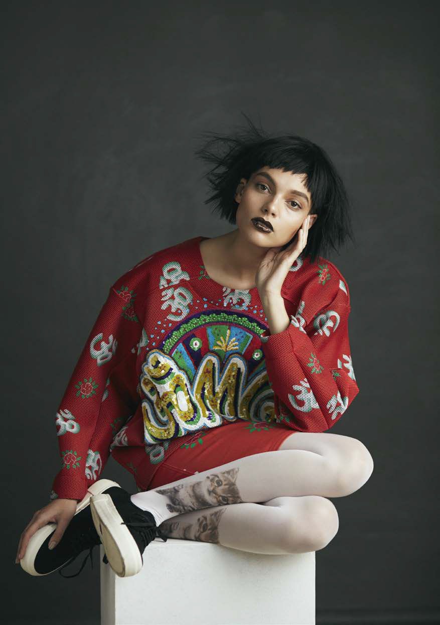 A young adult woman with dark lipstick and short black hair poses while seated on a white cube in a fashion editorial shot against a gray background.