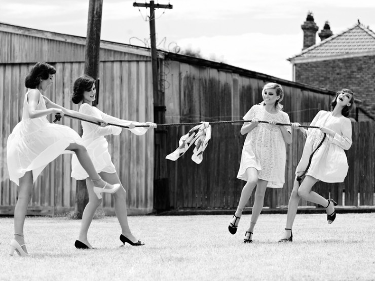 Four women in white dresses playfully engage in a tug-of-war on a grassy lawn, captured in a vintage-style black and white photograph.
