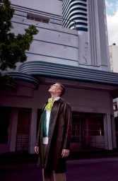 Man in a dark leather coat and bright yellow scarf looking up in front of an Art Deco building by Liz Ham.