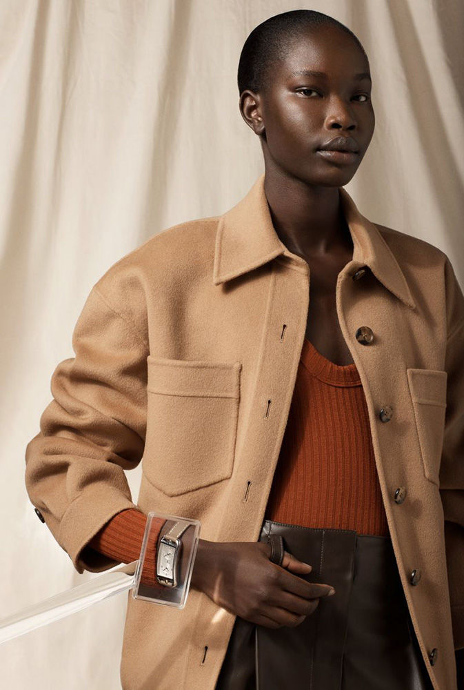 Close-up shot of a Black woman wearing a tan jacket and a silver wristwatch displayed in a clear plastic case, against a beige backdrop.
