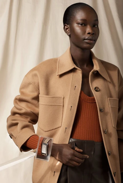 Close-up shot of a Black woman wearing a tan jacket and a silver wristwatch displayed in a clear plastic case, against a beige backdrop.