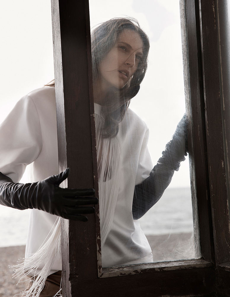 A woman in a white shirt and black gloves stands in front of a window. Outdoor setting, soft lighting.