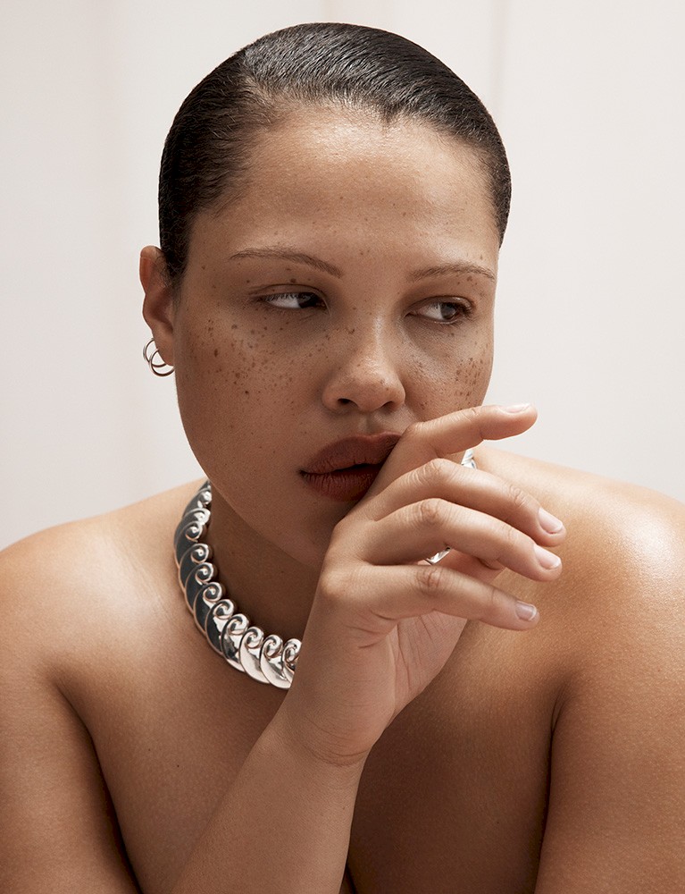 Close-up studio shot of an adult woman with freckles wearing a silver-tone necklace and posing with her hand near her mouth.