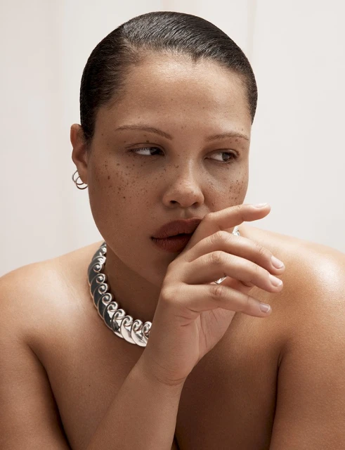 Close-up studio shot of an adult woman with freckles wearing a silver-tone necklace and posing with her hand near her mouth.