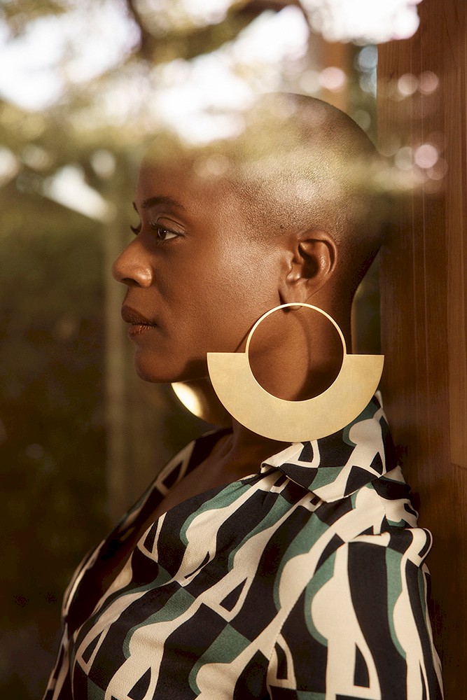 Portrait of a Black woman wearing large gold earrings and a patterned blouse, reflecting trees and sky from a window.