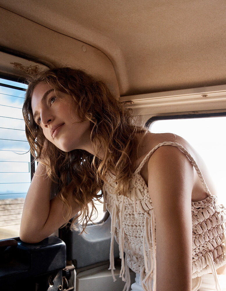 Portrait of a young adult woman with wavy brown hair sitting in a car, wearing a fringe-detail top, looking out the window.