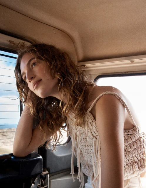 Portrait of a young adult woman with wavy brown hair sitting in a car, wearing a fringe-detail top, looking out the window.