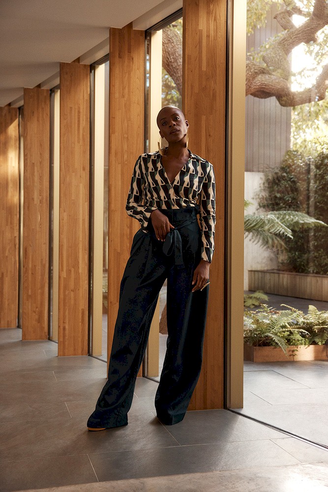 A Black female model poses indoors near a window, wearing a printed top with long sleeves and high-waisted, wide-leg pants with a tie belt.
