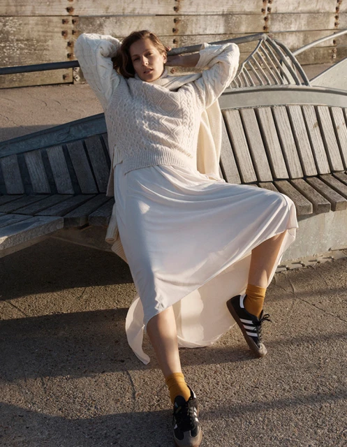 Adult female in a cable-knit sweater and long flowing skirt sitting on an outdoor bench in the afternoon sunlight with retro sneakers.