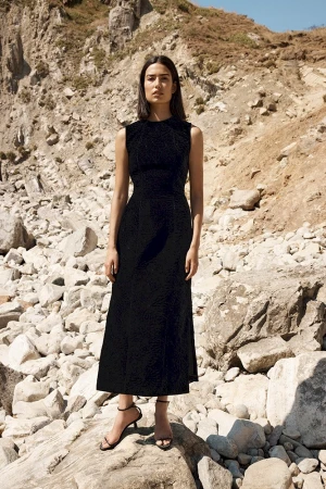 Woman in a black sleeveless midi dress standing on rocky terrain under natural sunlight