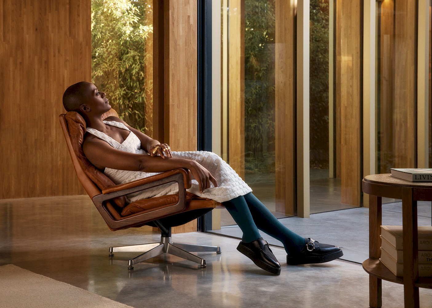 Woman rests in a brown leather chair in a minimalist space, wearing a white dress, green tights, and black loafers, bathed in natural light.