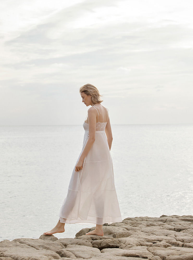 A woman in a white dress standing barefoot on rocks by the ocean, gazing down on a cloudy day, creating a serene fashion image.