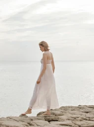 Woman in a long white dress standing on rocky coastal shore against an overcast sky and ocean by Amie Milne