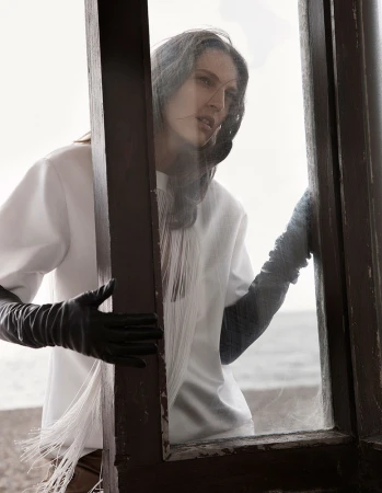 Woman in white top and black leather gloves looking out through a rustic wooden window frame near the beach