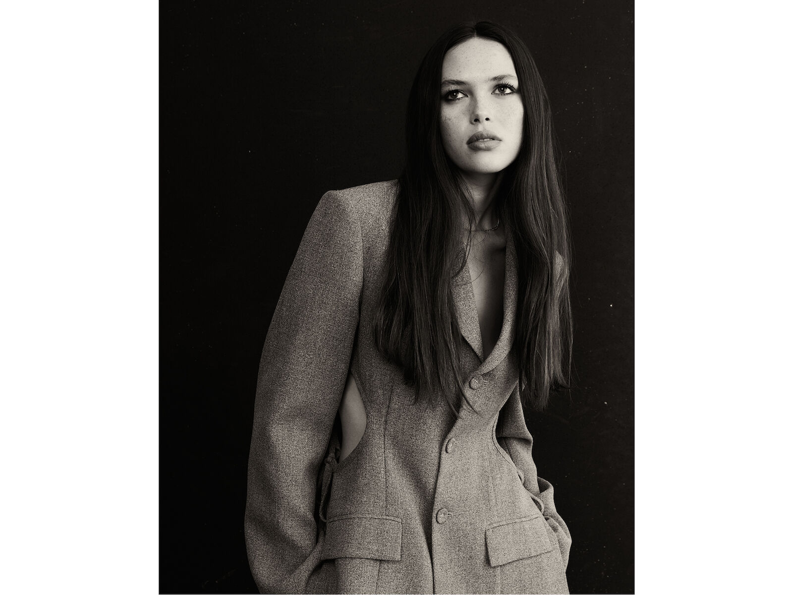 Monochrome portrait of a woman in a blazer against a dark backdrop, captured with a medium shot and eye-level angle.