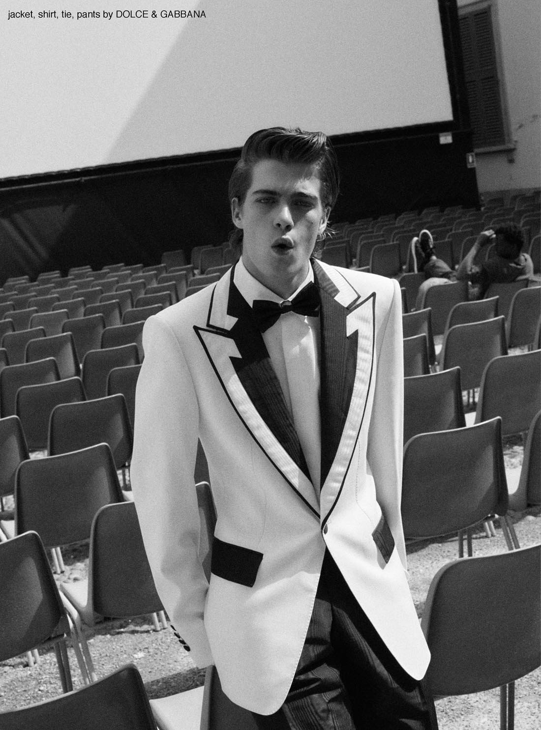Monochrome fashion shot of a young adult man in a tuxedo with a bow tie standing in an outdoor theater with rows of empty chairs.