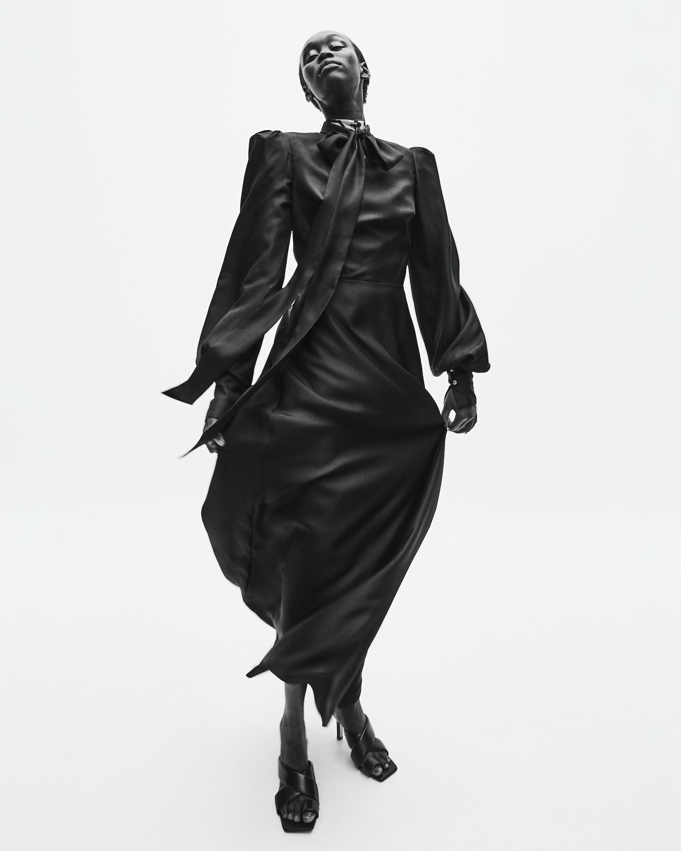 Elegant black and white fashion shot of a slim African woman in a long flowing black dress and heels, looking up in a studio setting.