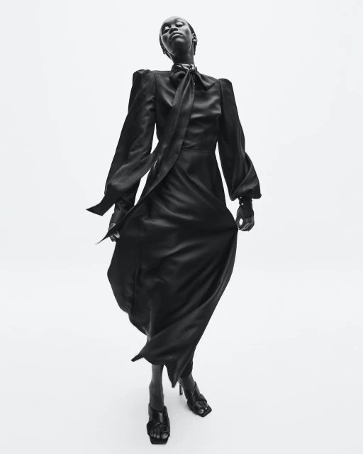 Elegant black and white fashion shot of a slim African woman in a long flowing black dress and heels, looking up in a studio setting.