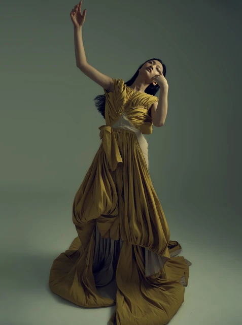 Elegant woman in mustard yellow gown strikes a pose in a studio. The floor-length dress features unique draping and flowing fabric.