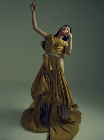Elegant woman in mustard yellow gown strikes a pose in a studio. The floor-length dress features unique draping and flowing fabric.