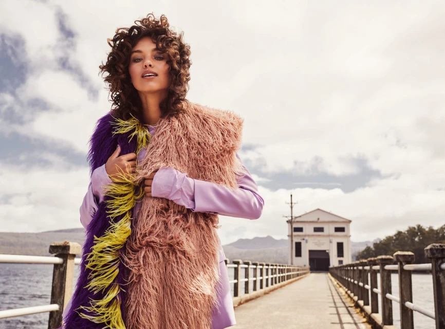 Fashion shot of a woman in a purple outfit and a multi-colored fur stole posing on a pier with a building and landscape in the background.