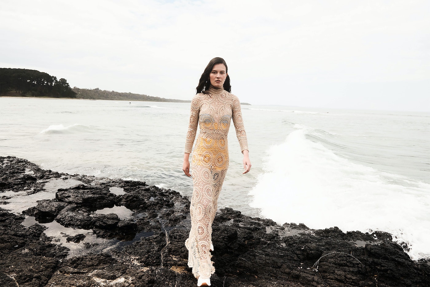 A slender woman with dark hair walks on rocky beach wearing a crocheted tan dress as waves crash on a cloudy day near wooded coast.