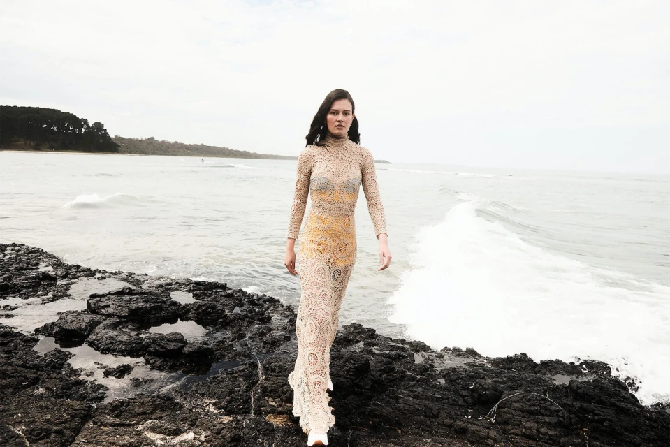A slender woman with dark hair walks on rocky beach wearing a crocheted tan dress as waves crash on a cloudy day near wooded coast.