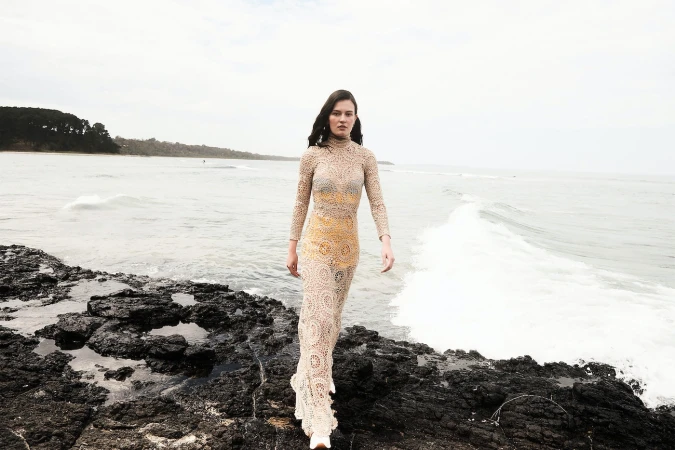 A slender woman with dark hair walks on rocky beach wearing a crocheted tan dress as waves crash on a cloudy day near wooded coast.