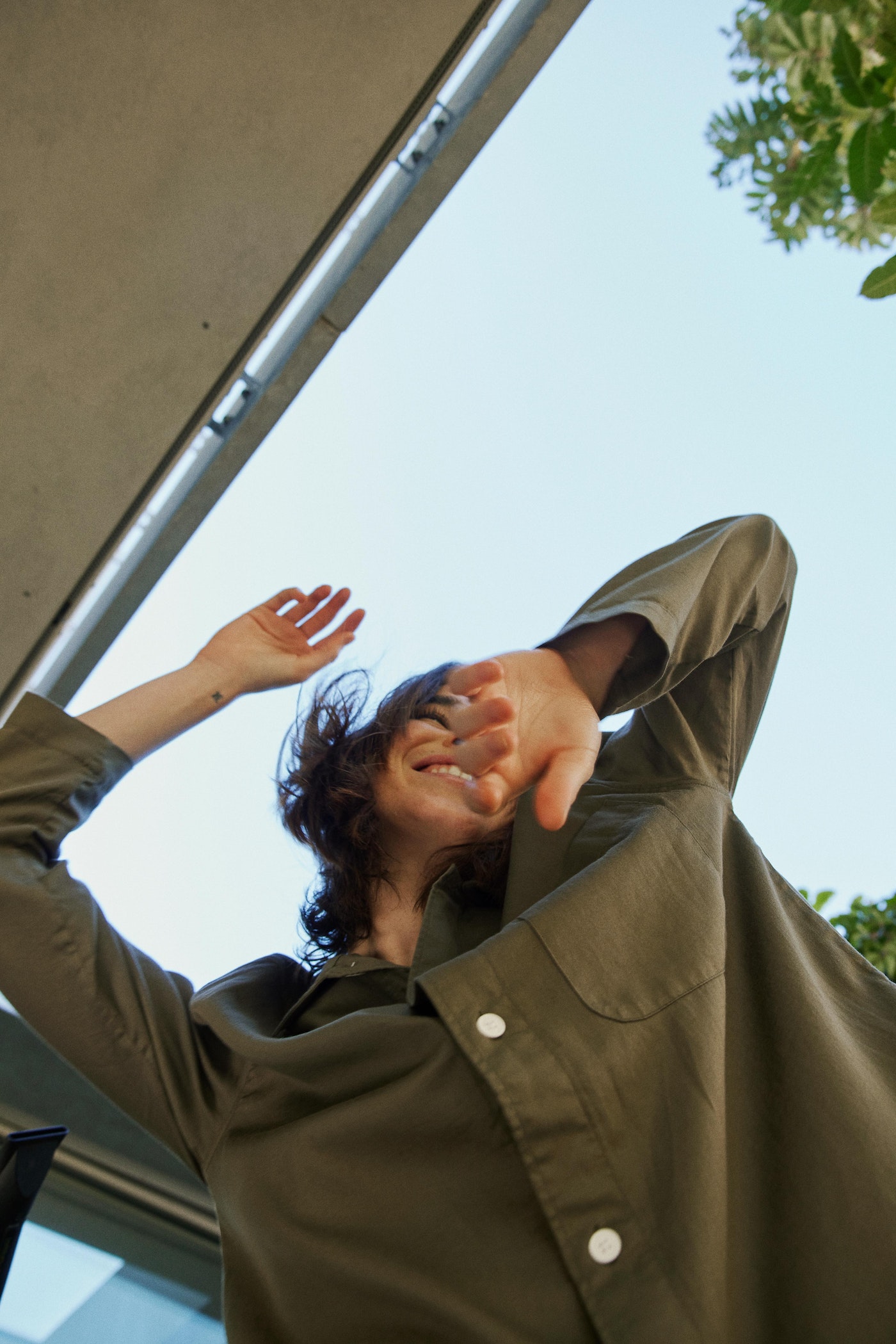 Woman in olive green shirt smiles with arms raised under modern architecture against a bright blue sky, capturing an upward perspective.