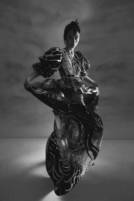 A woman in a flowing black-and-white patterned dress poses in a studio with a cloudy backdrop, artful lighting, and elegant fashion.