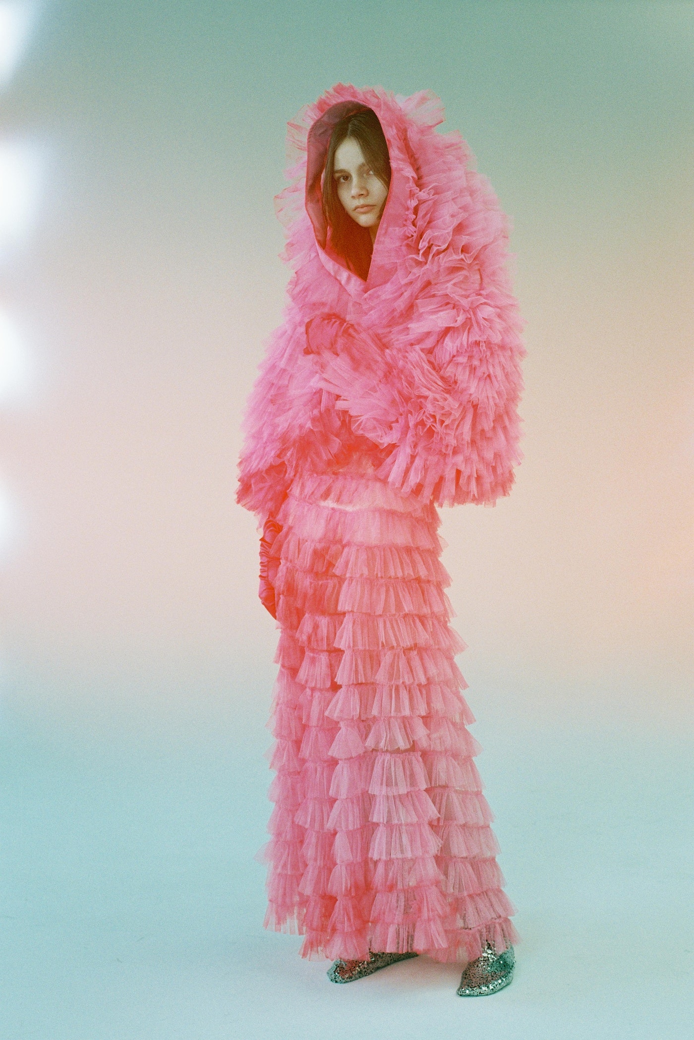 Young woman poses in a full-length shot, wearing a pink ruffled coat and skirt against a soft, diffused background, fashion studio setting.