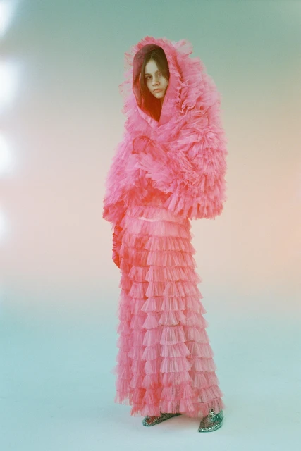 Young woman poses in a full-length shot, wearing a pink ruffled coat and skirt against a soft, diffused background, fashion studio setting.