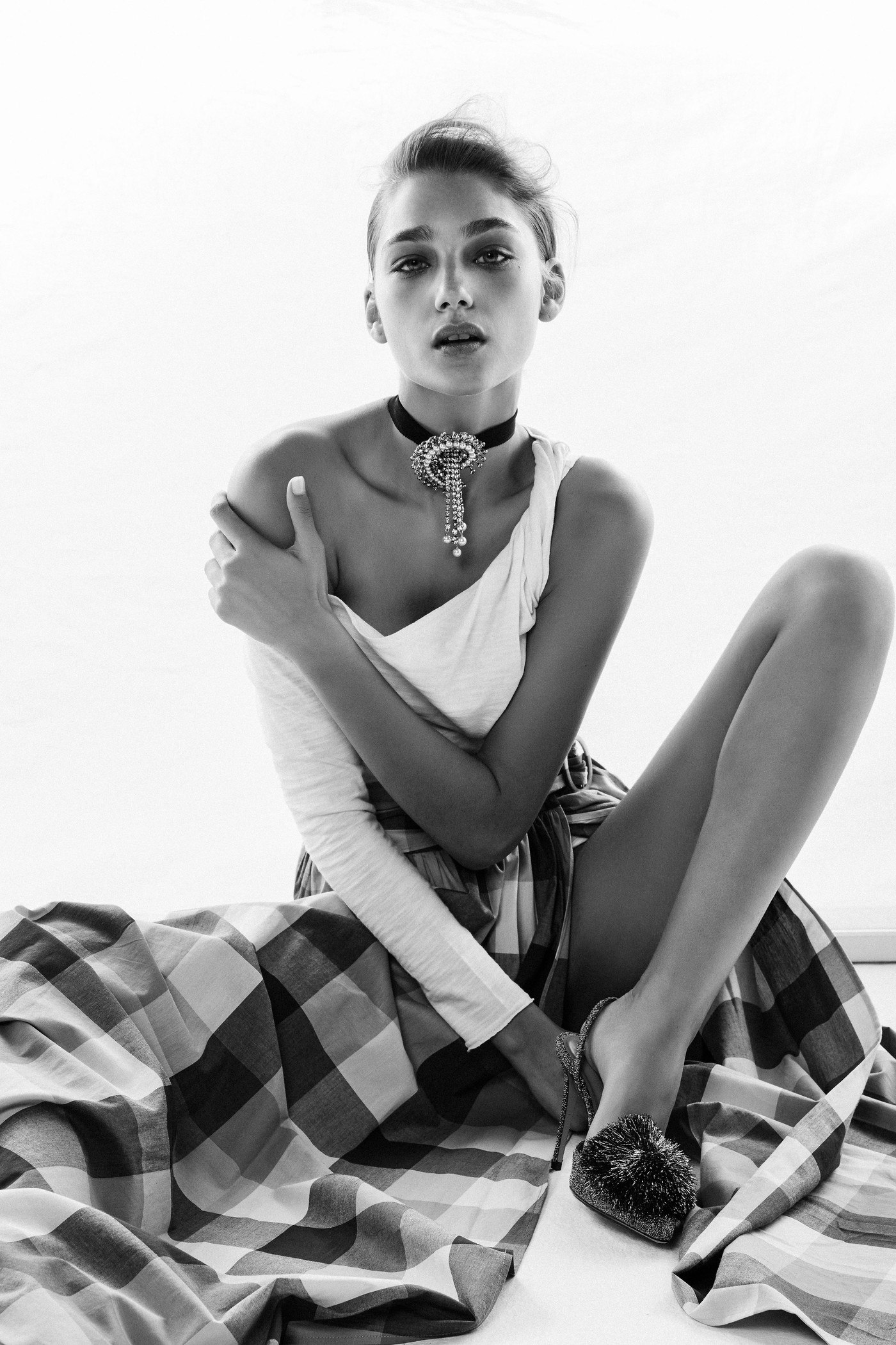 Black and white shot of a young adult woman in a white top and plaid skirt, sitting on the floor wearing a decorative choker necklace.