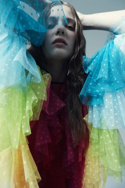Fashion portrait of a woman with her hands behind her head and wearing a colorful ruffled tulle dress with polka dots on a grey background.